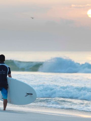 Surfer steht mit Brett am Strand und schaut auf die untergehende Sonne.