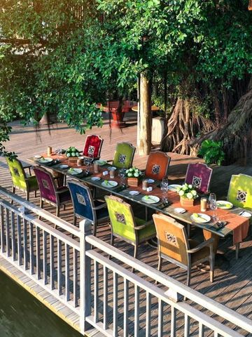 Außenbereich mit langem Esstisch und bunten Stühlen direkt am Flussufer unter einem Baum