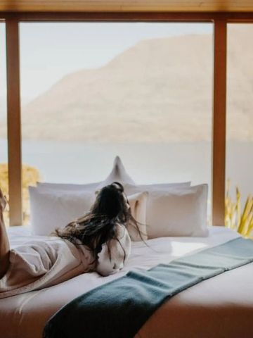 Frau in Bademantel auf Bett liegend mit Seeblick durch Panoramafenster