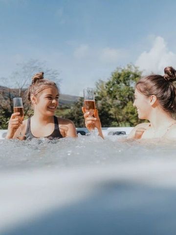 Zwei Personen stoßen mit Getränken im Whirlpool an, umgeben von Natur.