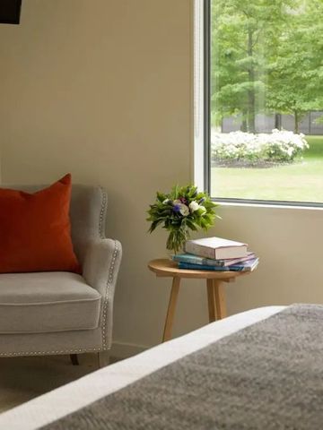 Gemütliche Sitzecke mit Sessel, rotem Kissen und Tisch vor einem großen Fenster.