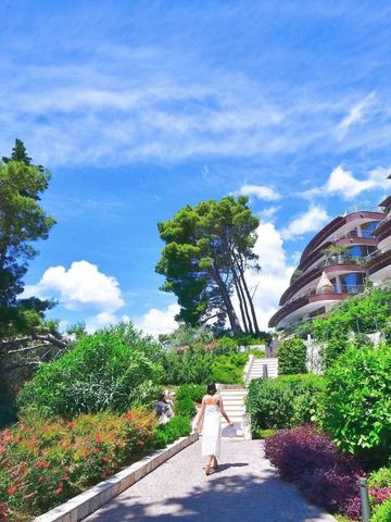 Frau in weißem Kleid spaziert durch grünen Gartenbereich vor dem modernen Hotelgebäude unter strahlend blauem Himmel