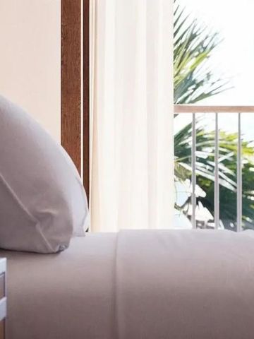 Bett mit Kissen an einem Fenster mit Blick auf Palmen und das Meer