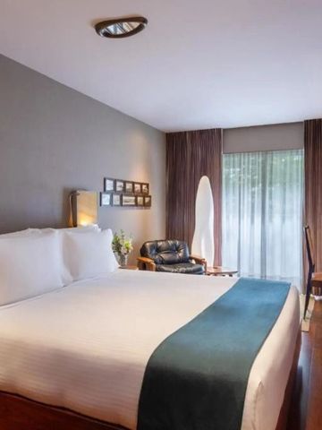 Modernes Schlafzimmer mit großem Bett, Sesseln und Tisch. Helle Vorhänge am Fenster.