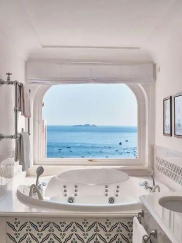 Whirlpool mit Meerblick durch großes Fenster in einem hellen Badezimmer.