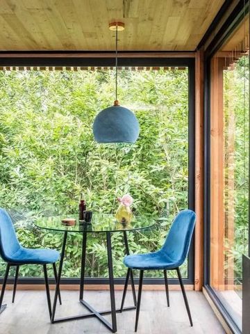 Essbereich mit Glaswänden, Waldblick, Glasplatte und blauen Stühlen.