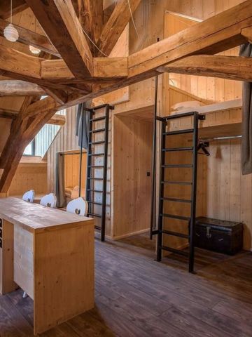 Gemütliches Holzzimmer mit rustikalen Balken, Leitern zu Hochbetten und einem langen Tisch.