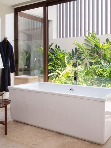 Freistehende Badewanne vor bodentiefen Fenstern mit Gartenblick.