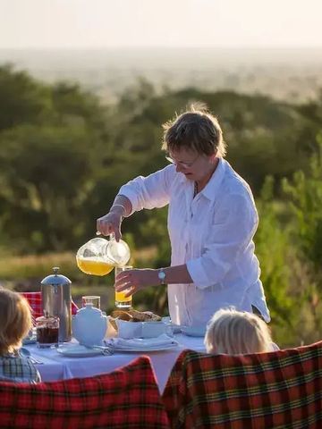 Frau serviert Frühstück an einem Tisch im Freien mit Kindern in der Serengeti