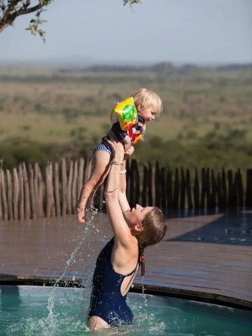 Frau hebt Kind im Pool mit afrikanischer Savannenlandschaft im Hintergrund