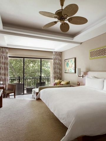 Helles Hotelzimmer mit großem Bett, Deckenventilator und Balkonblick ins Grüne.