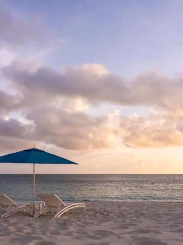 Zwei Liegestühle unter einem blauen Sonnenschirm am Sandstrand bei Sonnenuntergang