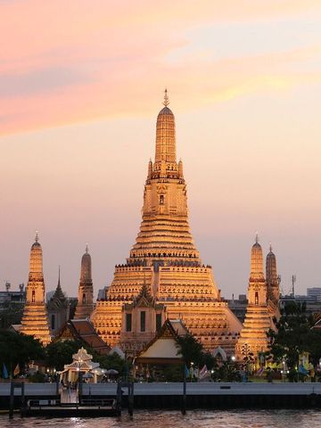 Der Wat Arun erstrahlt im warmen Licht des Sonnenuntergangs, seine filigranen Türme spiegeln sich sanft im ruhigen Fluss und schaffen eine magische, friedvolle Atmosphäre.