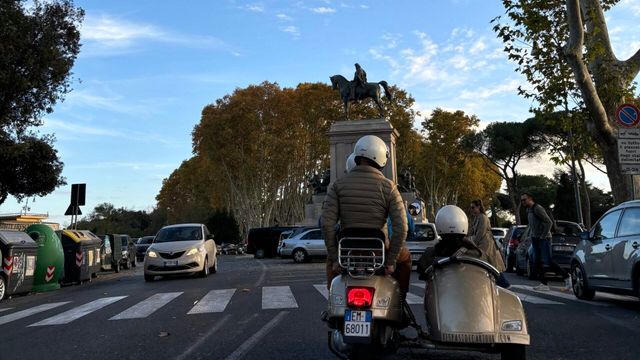 Zwei Personen auf einem Roller mit Beiwagen vor einer Reiterstatue an einer belebten Kreuzung.