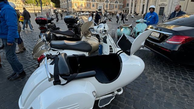 Parkplatz mit Motorrollern und Autos auf einem alten Kopfsteinpflaster in einer Stadt.