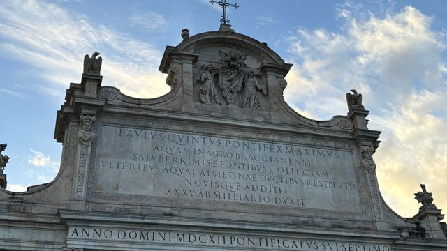 Detail der oberen Fassade eines Bauwerks mit lateinischer Inschrift und einer Statue.