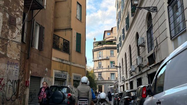 Schmale Gasse mit geparkten Fahrzeugen und zwei Menschen auf Rollern in einer Stadt.