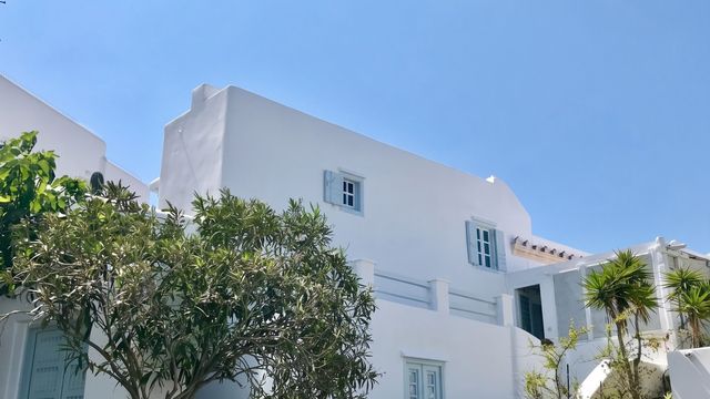 Weiß getünchtes Gebäude im kykladischen Stil mit hellblauen Fensterläden und grünen Pflanzen.