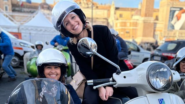 Zwei lachende Frauen mit Helmen auf einem weißen Motorroller vor historischer Architektur.