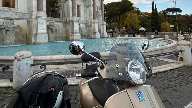 Vespa vor einem historischen Brunnen mit neoklassizistischen Details und Bäumen.