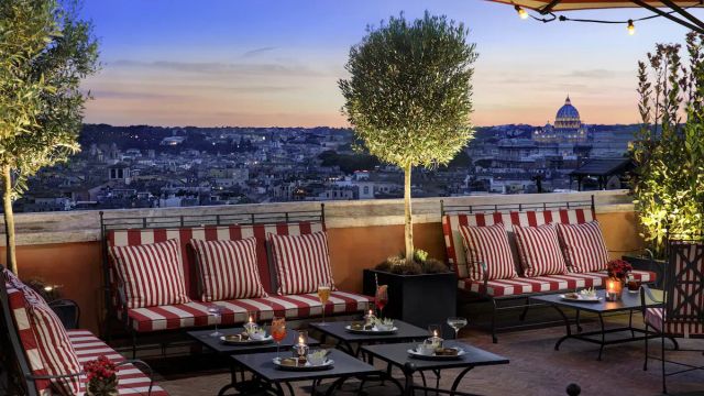 Elegante Dachterrasse des Rocco Forte Hotel de la Ville in Rom mit gestreiften Sitzpolstern, Cocktailtischen und Blick auf die Stadt sowie den Petersdom bei Sonnenuntergang.