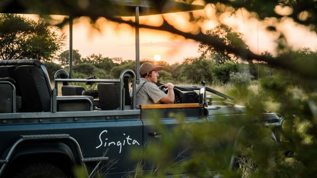 Ein Guide steuert ein Safari-Fahrzeug bei Sonnenuntergang durch die bewachsene Savannenlandschaft.