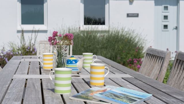 Holztisch auf der Terrasse mit bunten Tassen, Blumenvase und Magazinen vor weißer Fassade