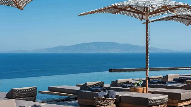 Infinity-Pool mit Liegen und Sonnenschirmen vor malerischem Meerblick und Hügeln am Horizont