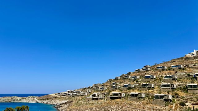 Häuser reihen sich an einem Hügel entlang mit blauem Himmel und Meer im Hintergrund.