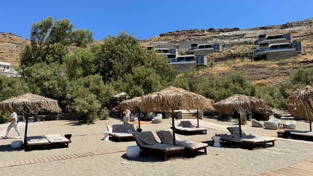 Liegen unter Strohschirmen am Strand des One&Only Kéa Island, dahinter die Villen am Hang