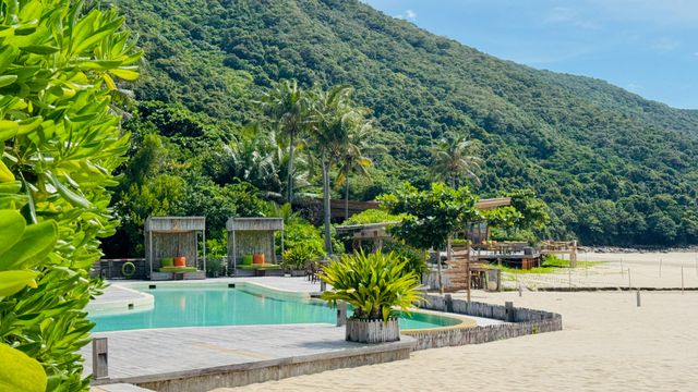 Strandresort mit Pool vor üppigen grünen Hügeln und weißem Sandstrand.