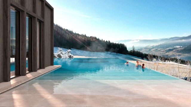 Infinity-Außenpool des AKI Family Resort PLOSE mit Blick auf verschneite Wiesen und ein weites Tal in Südtirol, Menschen baden im warmen Wasser vor moderner Holzarchitektur.