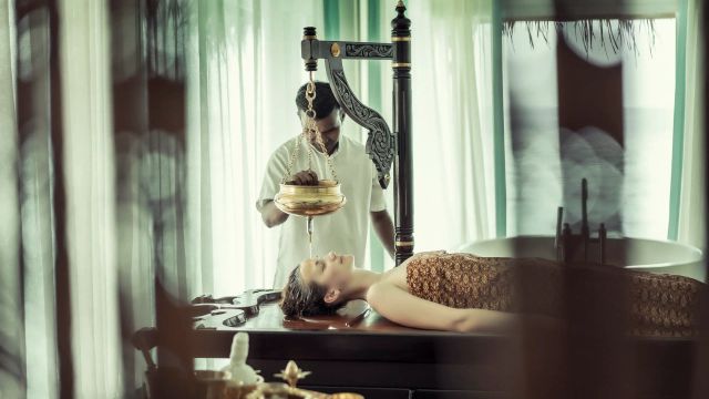 Ayurvedische Shirodhara-Behandlung im Spa des Anantara Kihavah Maldives Villas, bei der warmes Öl auf die Stirn einer entspannt liegenden Frau gegossen wird.