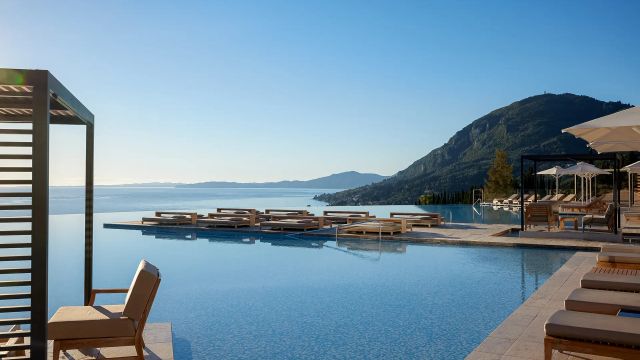 Infinity-Pool des Angsana Corfu Resorts mit eleganten Liegeflächen und Blick über das glitzernde Ionische Meer, eingerahmt von einem bewaldeten Berghang und unter wolkenlosem, strahlend blauem Himmel – eine Szenerie voller Ruhe und Luxus.