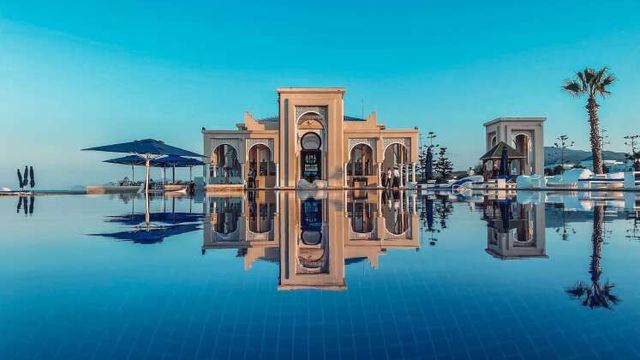 Majestätisches Gebäude im maurischen Stil im Banyan Tree Tamouda Bay, gespiegelt in einem ruhigen Infinity-Pool, umgeben von Palmen und luxuriöser Resort-Atmosphäre bei strahlend blauem Himmel.