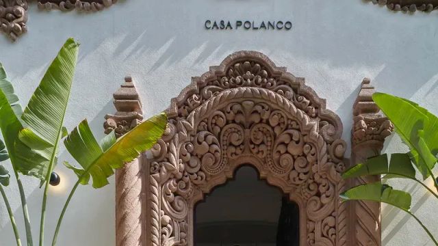 Prächtig verzierter steinerner Eingangsbereich des Boutiquehotels Casa Polanco in Mexiko-Stadt, mit kunstvollen Ornamenten im Kolonialstil und flankiert von großen tropischen Pflanzen vor einer weiß verputzten Fassade.