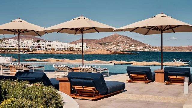 Poolterrasse des Cosme, a Luxury Collection Resort, Paros mit Sonnenliegen, Sonnenschirmen und Blick auf Meer, weiße Häuser und Hügelkulisse.
