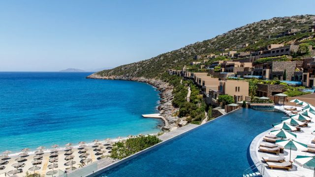 Blick auf den Infinity-Pool des Daios Cove Luxury Resort & Villas auf Kreta mit Sonnenliegen, türkisblauem Meer, Privatbucht und terrassenförmig.