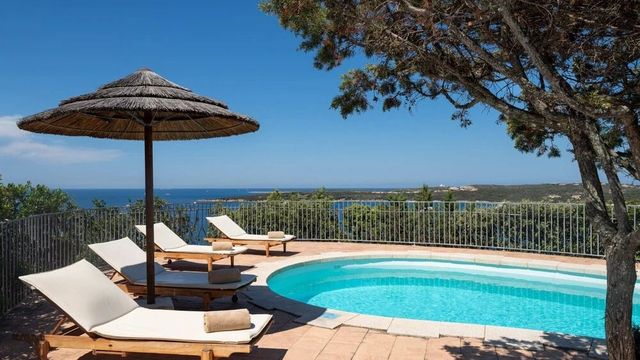 Kleiner Pool mit Sonnenliegen und Strohdach-Sonnenschirm im Hotel Pitrizza auf Sardinien, umgeben von Bäumen und mit weitem Blick auf die grüne Küste und das blaue Mittelmeer.