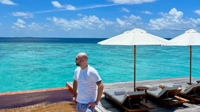 Timo teht in einem Pool mit Mosaikfliesen auf einer Holzterrasse am türkisblauen Meer der Malediven, umgeben von Sonnenliegen und weißen Sonnenschirmen.