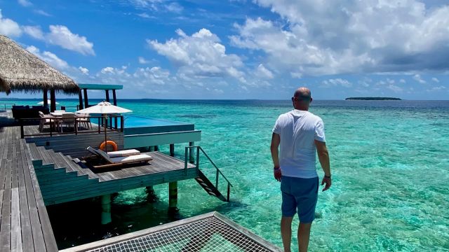Timo steht auf einer Netzliege über dem türkisblauen Meer auf den Malediven, umgeben von Wasser-Villen und strahlend blauem Himmel.