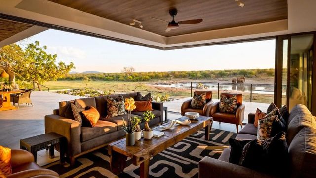 Offene Lounge der Jamala Madikwe Royal Safari Lodge mit eleganten Sofas, Holztisch, afrikanischem Dekor und atemberaubendem Blick auf ein Wasserloch mit Elefanten – luxuriöses Safari-Ambiente in freier Natur.