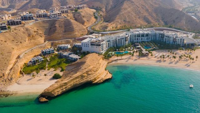 Luftaufnahme des Jumeirah Muscat Bay in Oman mit direkter Strandlage, türkisfarbenem Wasser, Poollandschaft, eleganter Architektur und umgeben von einer eindrucksvollen Bergkulisse.