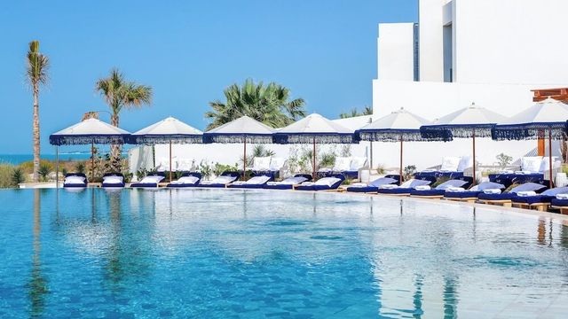 Poolbereich des Jumeirah Saadiyat Island Resorts mit weißen Sonnenschirmen, blauen Liegen, Palmen und Meerblick