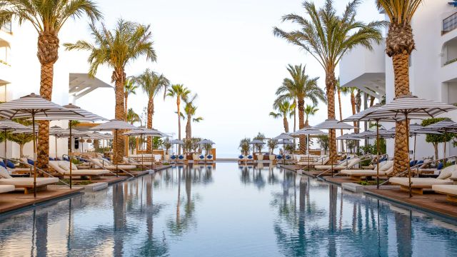 Stilvoller Poolbereich des METT Hotel & Beach Resort mit Palmen, gepolsterten Sonnenliegen, gestreiften Sonnenschirmen und Blick Richtung Meer, eingerahmt von weißer moderner Architektur.