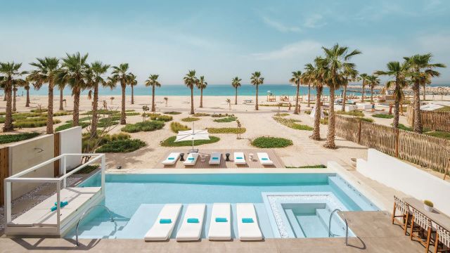 Blick auf den Poolbereich einer privaten Villa im Nikki Beach Resort Dubai mit modernen Sonnenliegen, Palmen, Sandstrand und türkisblauem Meer im Hintergrund – luxuriöse Strandatmosphäre.