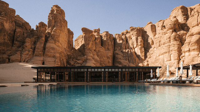Luxuriöser Pool mit türkisblauem Wasser vor einem flachen, modernen Gebäude, eingerahmt von eindrucksvollen rötlich-gelben Sandsteinfelsen unter klarem, blauem Himmel. 