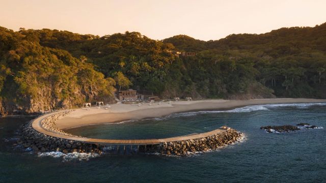Luftaufnahme eines luxuriösen Strandresorts mit halbkreisförmiger Mole, weißem Sandstrand und dichter tropischer Vegetation im Hintergrund.