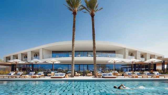Moderner Poolbereich des One&Only Aesthesis in Griechenland mit eleganter Architektur, zwei hohen Palmen, stilvollen Sonnenliegen und einem Schwimmer im türkisfarbenen Wasser vor klarblauem Himmel.