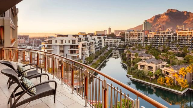 Aussicht vom Balkon des One&Only Cape Town mit Blick auf moderne Wohnanlagen, eine ruhige Wasserstraße und den Tafelberg im Hintergrund bei Sonnenuntergang.